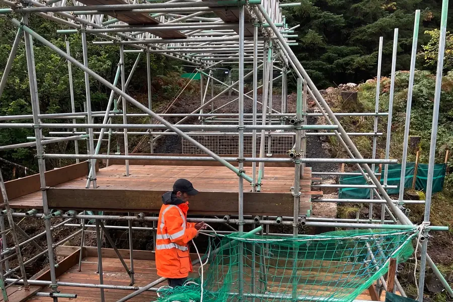 Scaffolder in hi-vis PPE fitting safety netting to a scaffold structure on a rural access project